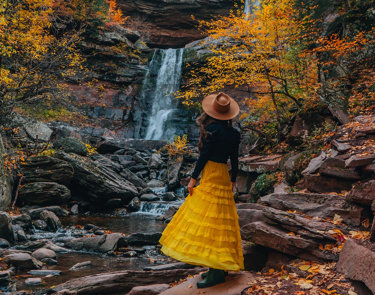Woman Standing in Yellow Dress Boots a sweater and Hat in front of waterfall with fall foliage